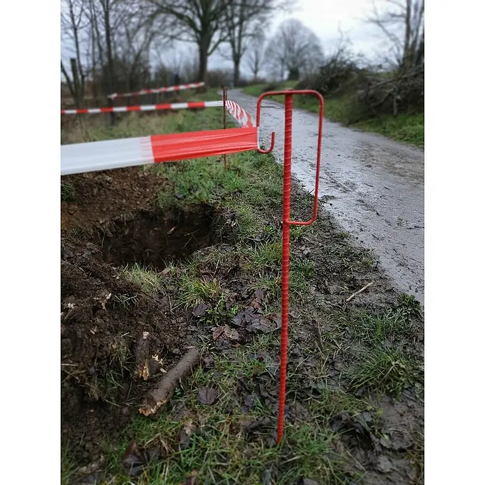 Absperrleinenhalter - 1,25 m, Sicherheitsbügel, Rot, Geschmiedet - Ein roter, stabförmiger Erdanker mit Haken für Absperrband steht vor einem Loch, um vor Gefahren zu warnen.