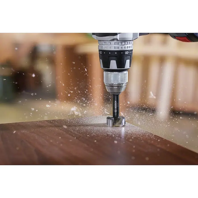 Bosch Forstnerbohrer-Set - 5 -tlg. - Nahaufnahme einer Bohrmaschine mit Bosch-Aufsatz, die ein Loch in ein dunkles Holzbrett bohrt, wobei Holzspäne umherfliegen.