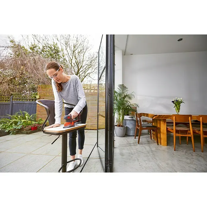 Black+Decker 18V PowerConnect Lijadora delta de batería BDCDS18N-XJ - 18 V, Iones de litio, Sin batería, 24.000 r.p.m. - Mujer lijando un taburete de bar con una lijadora BLACK+DECKER en un patio que se abre al salón.