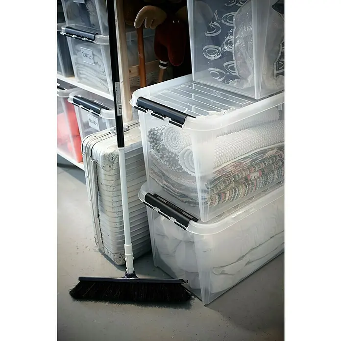 SmartStore Aufbewahrungsbox Classic 70 - L x B x H: 72 x 40 x 39 cm, Kunststoff, Transparent - Nahaufnahme von einer schwarzen Kehrbürste, einem silberfarbenen Koffer und transparenten Aufbewahrungsboxen mit Decken und Textilien.