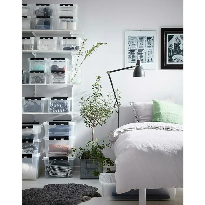 SmartStore Aufbewahrungsbox Classic 8 - L x B x H: 34 x 25 x 16 cm, Kunststoff, Transparent - Das Bild zeigt ein Schlafzimmer mit einem Bett und einer Wand mit transparenten Aufbewahrungsboxen, die Kleidung und Accessoires enthalten. Eine Stehlampe ist auch zu sehen.