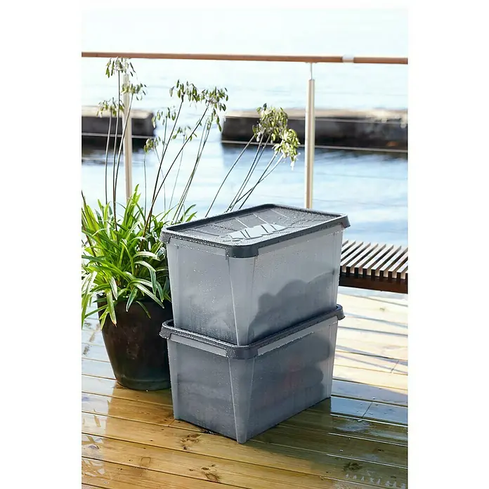 SmartStore Aufbewahrungsbox Dry 55 - 55 l, Lebensmittelecht, Wasserdicht, Stapelbar - Zwei graue Kunststoffboxen mit Deckel stehen auf einer nassen Holzterrasse, daneben ein Topf mit grünen Pflanzen.