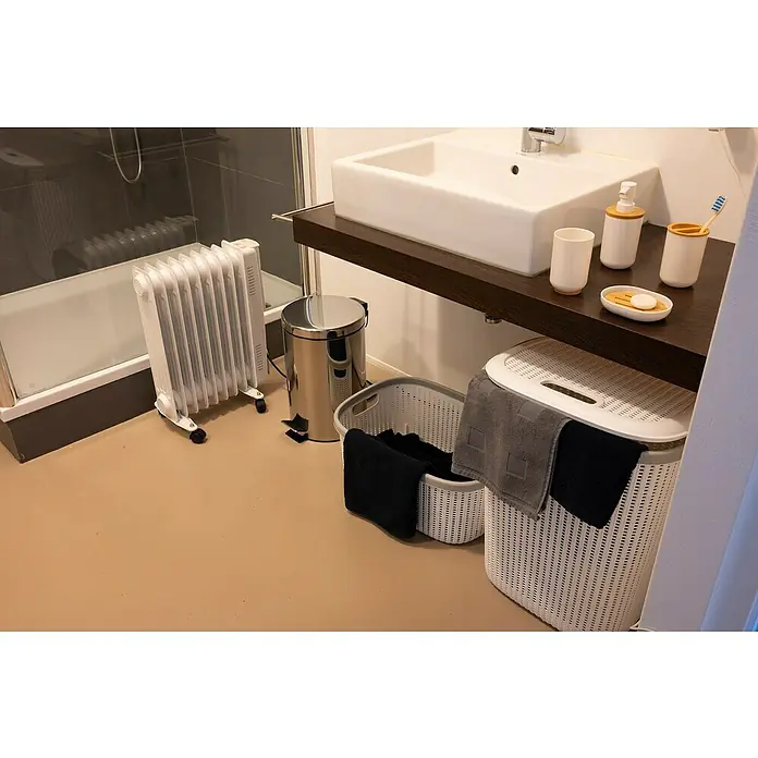 Radiador de aceite Alpina - 23,5 x 55 x 40 cm - Vista del baño con radiador, cubo de basura, cestos de ropa y lavabo. La habitación está equipada con elementos funcionales.
