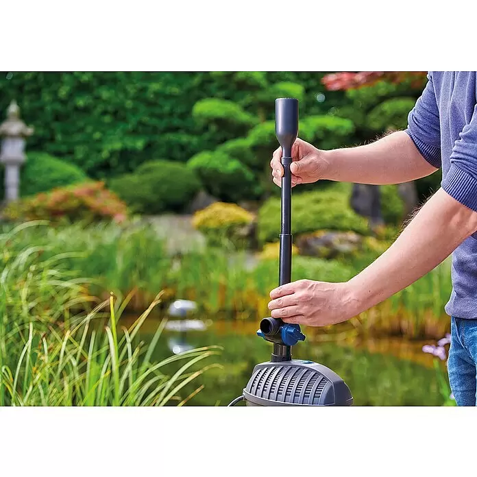 Oase Wasserspielpumpe Aquarius Set 2000 E - 18 W, Fördermenge pro Std.: 2.100 l - Hände montieren eine dunkelgraue und blaue Teichpumpe mit Fontänenaufsatz in einem üppigen Garten an einem Teich.