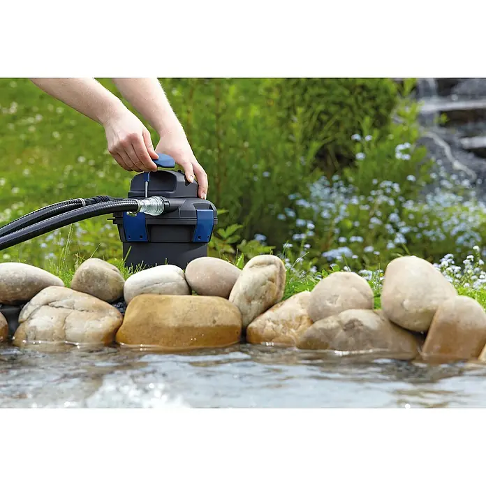Oase Druckfilter BioPress Set 4000 - 25 W, Teichgröße: 4 m³ - Ein Teichfilter wird an einem Teich mit Felsen installiert. Der Filter ist schwarz mit blauen Akzenten und ist mit Schläuchen verbunden.