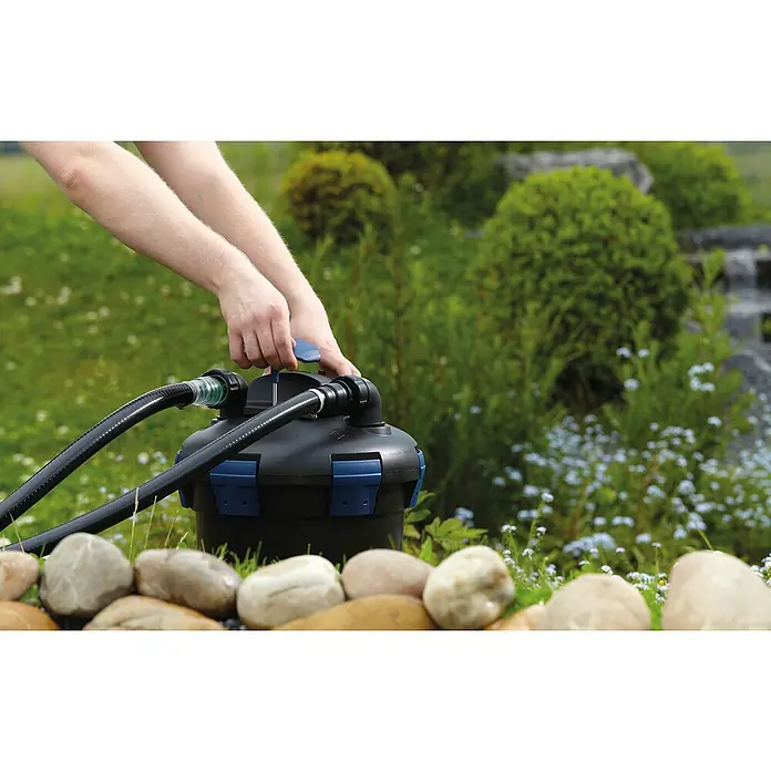 Oase Druckfilter BioPress Set 6000 - 40 W, Teichgröße: 6 m³ - Ein schwarzer Teichfilter mit blauen Akzenten wird von Händen im Garten bedient. Schläuche sind angeschlossen und Steine liegen im Vordergrund.