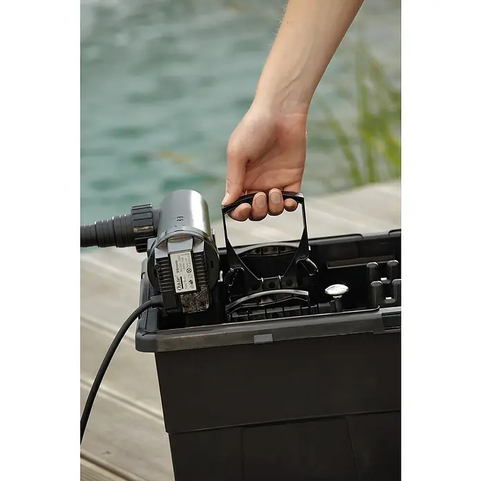 Oase BioSmart Durchlauffilter Set 14000 - 70 W, Teichgröße: 14 m³ - Eine Person trägt einen Oase Teichfilter in einer schwarzen Box, umgeben von einer Holzterrasse und Wasser im Hintergrund.