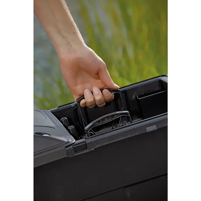 Oase BioSmart Durchlauffilter Set 14000 - 70 W, Teichgröße: 14 m³ - Eine Hand hält den Griff eines schwarzen Angelkastens vor einem unscharfen, grün-braunen Hintergrund.