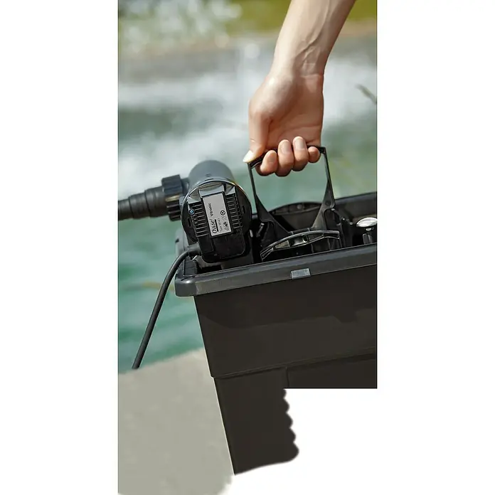 Oase BioSmart Durchlauffilter-Set 5000 - 25 W, Teichgröße: 5 m³ - Eine Person trägt eine schwarze Oase-Box, die einen Wasserfilter enthält, vor einem verschwommenen Wasserhintergrund.