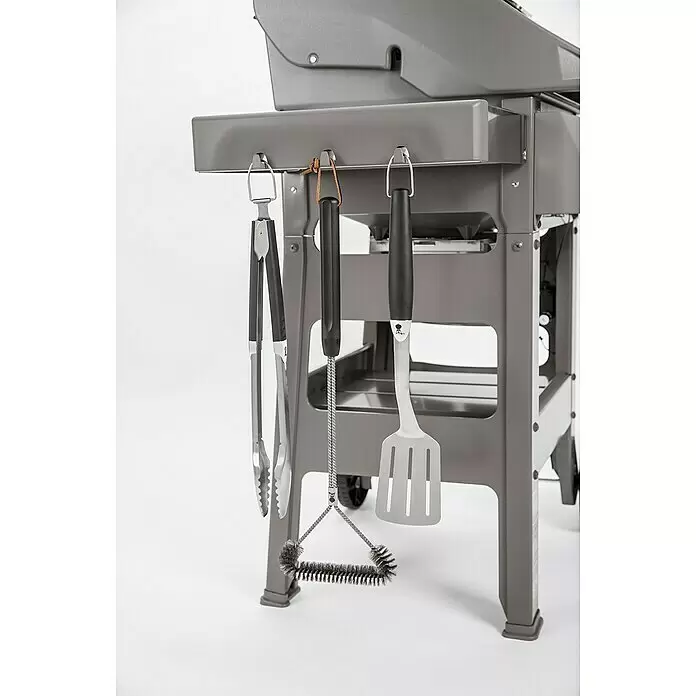 Weber Barbacoa de gas Spirit II E-310 + 7 accesorios - Superficie de la parrilla (An x Pr): 60 x 46 cm, 8,7 kW, Número de quemadores: 3 - Tres utensilios de parrilla – pinzas, cepillo y espátula de acero inoxidable – colgando del lateral de una parrilla gris.