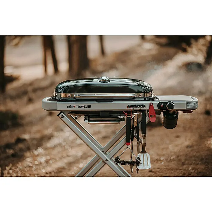 Weber Barbacoa de gas Traveler - Superficie de la parrilla (An x Pr): 64 x 35 cm, 3,8 kW, Número de quemadores: 1 - Primer plano de una parrilla Weber Traveler al aire libre, con utensilios de cocina. La parrilla está lista para una deliciosa experiencia al aire libre.