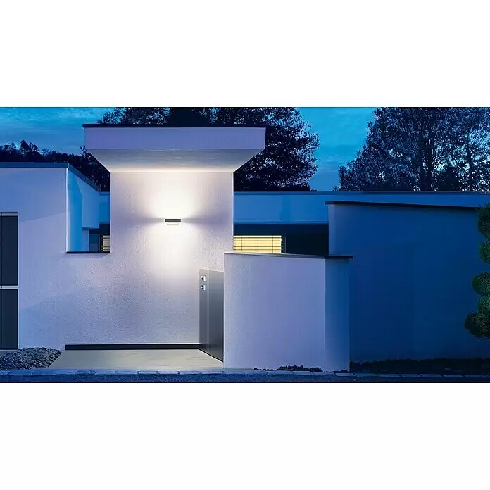 Steinel LED-Sensor-Außenwandleuchte L 810 SC anthrazit - L x B x H: 14,6 x 23 x 9,1 cm - Eine elegante, dunkle Außenwandleuchte spendet oben-unten Licht an einem modernen weißen Gebäude bei Dämmerung.