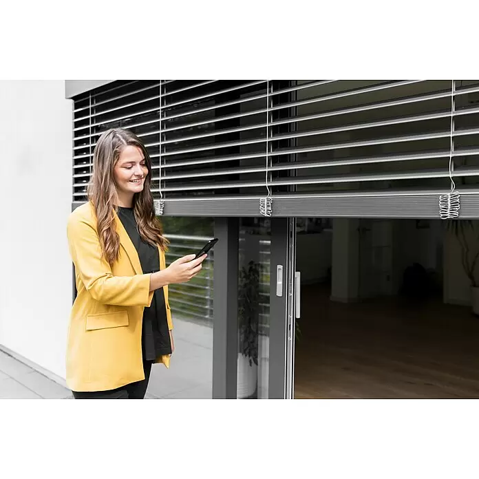 Schellenberg Smart Home Interruptor de recepción por radio - 80 x 80 x 40 mm, Blanco, Montaje enrasado - Una mujer sonríe mientras controla persianas venecianas exteriores grises con un smartphone.