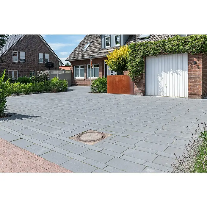 Diephaus Pflasterstein LincolnBasalt, 60 x 30 x 8 cm, Rechteckig - Ein gepflasterter Hof mit einem Kanaldeckel im Vordergrund und zwei Backsteinhäusern im Hintergrund. Das Pflaster ist grau und rechteckig.