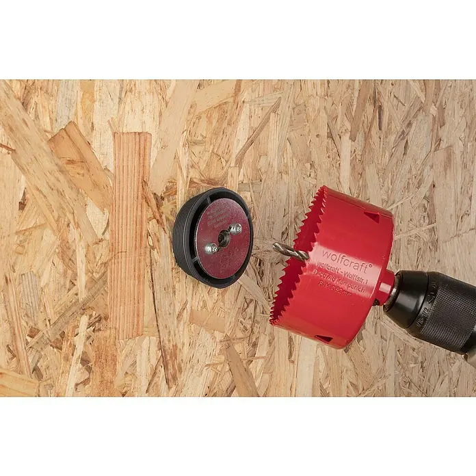 Wolfcraft Aufbohrhilfe-Set - 7 -tlg., Passend für: Lochsägen ab Ø 60 mm - Nahaufnahme einer roten Lochsäge der Marke Wolfcraft, die sich an einem Holzkasten befindet, um ein Loch zu bohren.