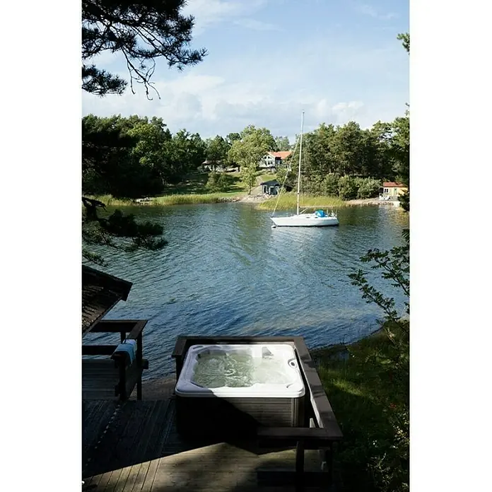 Camargue Außen-Whirlpool Emotion - 213 x 213 x 87 cm, Grau/Weiß, Max. Personenzahl: 6 - Entspannen Sie im Whirlpool mit Seeblick. Der Whirlpool ist auf einer Holzterrasse mit Blick auf einen ruhigen See mit einem Segelboot platziert.
