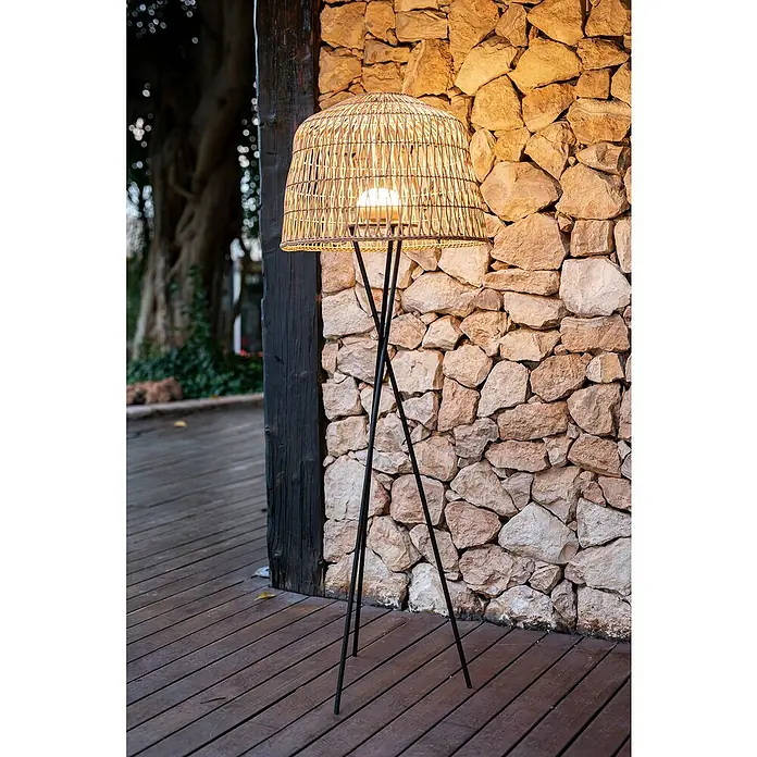 New Garden Außenstehleuchte Amalfi - L x B x H: 49 x 49 x 148 cm - Eine moderne Stehlampe mit einem Lampenschirm aus Rattan und einem schlanken, schwarzen Metallständer vor einer rustikalen Steinmauer.