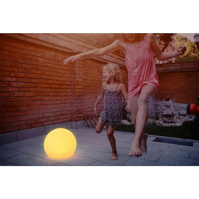 New Garden Solar-Dekoleuchte Buly - Durchmesser: 50 cm, IP65 - Ein leuchtender Globus erhellt das Bild, während zwei Kinder im Garten unter einem Rasensprenger spielen. Die Szene strahlt Freude aus.