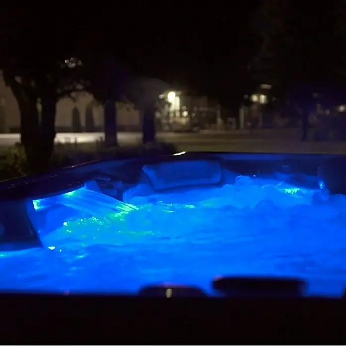 Camargue Außen-Whirlpool Serenity - 213 x 213 x 91 cm, Grau/Weiß, Max. Personenzahl: 6 - Nahaufnahme eines Whirlpools bei Nacht, beleuchtet mit blauem und grünem Licht, der Entspannung und Luxus verspricht.