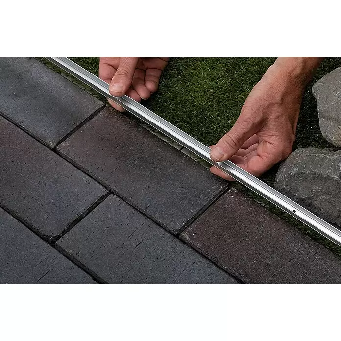 Paulmann Plug & Shine Montageschiene für Neon Stripe - Länge: 1 m - Hände platzieren eine silberne Aluminium-Gartenbegrenzung neben dunklen Pflastersteinen.