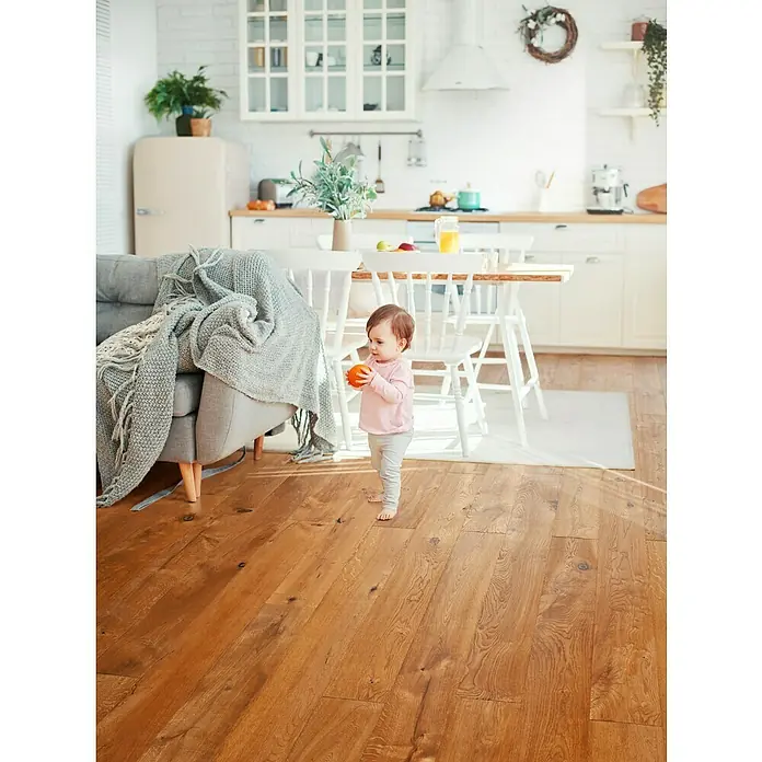 Parkett - Eiche Byzanz, Landhausdiele, 1.900 x 190 x 14 mm - Kleines Mädchen mit orangefarbenem Spielzeugball in einem hellen Raum mit Holzboden, Sofa und Esstisch. Blick auf die Küche im Hintergrund.