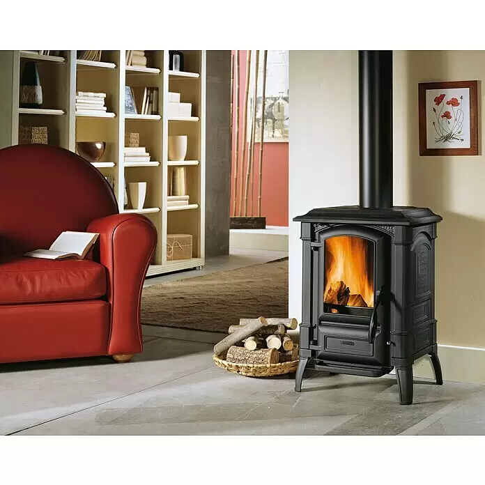 La Nordica Kaminofen Giulietta X - 8,2 kW, Raumheizvermögen: 189 m³, Material Verkleidung: Stahl, Schwarz/Anthrazit - Schwarzer Holzofen mit brennendem Feuer im Wohnzimmer neben rotem Sessel und Holzkorb.