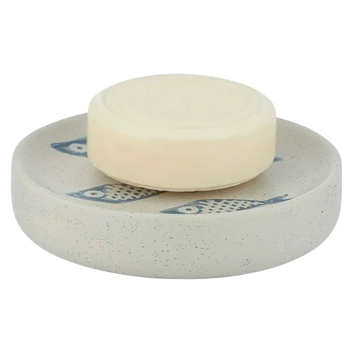 Wenko Seifenschale Aquamarin - Keramik, Beige - Ein rundes Seifenstück liegt auf einer Seifenschale mit blauem Fischmuster vor einem schlichten, schwarzen Hintergrund.