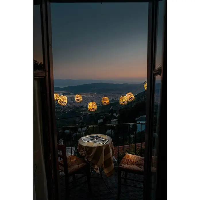 New Garden Solar-Lichterkette Aurora - Länge: 8 m, IP44 - Romantischer Balkonblick mit beleuchteten Laternen. Ein Tisch mit zwei Stühlen lädt zum Verweilen ein, während die Abenddämmerung die Szenerie erleuchtet.