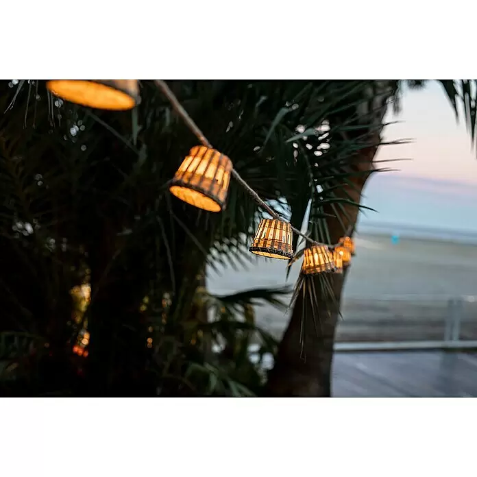 New Garden Solar-Lichterkette Aurora - Länge: 8 m, IP44 - Lichterkette mit geflochtenen Lampenschirmen an einer Palme vor Strandkulisse bei Dämmerung.