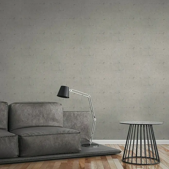 A.S. Création Elements Vliestapete Steinfliese - Grau/Beige, Betonoptik, 10,05 x 0,53 m - Nahaufnahme eines stilvollen Interieurs mit einem grauen Sofa, einer modernen Lampe und einem schwarzen Beistelltisch vor einer Betonwand.