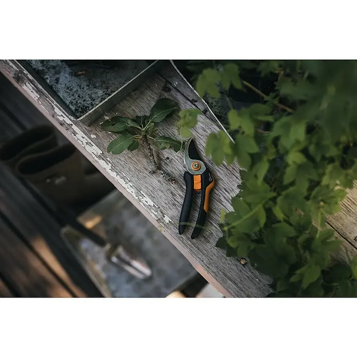 Fiskars Solid Gartenschere P321Schneidsystem: Bypass, Max. Aststärke: 20 mm - Gartenschere und ein kleiner Ast liegen auf einem verwitterten Holztisch, umgeben von üppigem Grün, was auf Gartenarbeiten hindeutet.