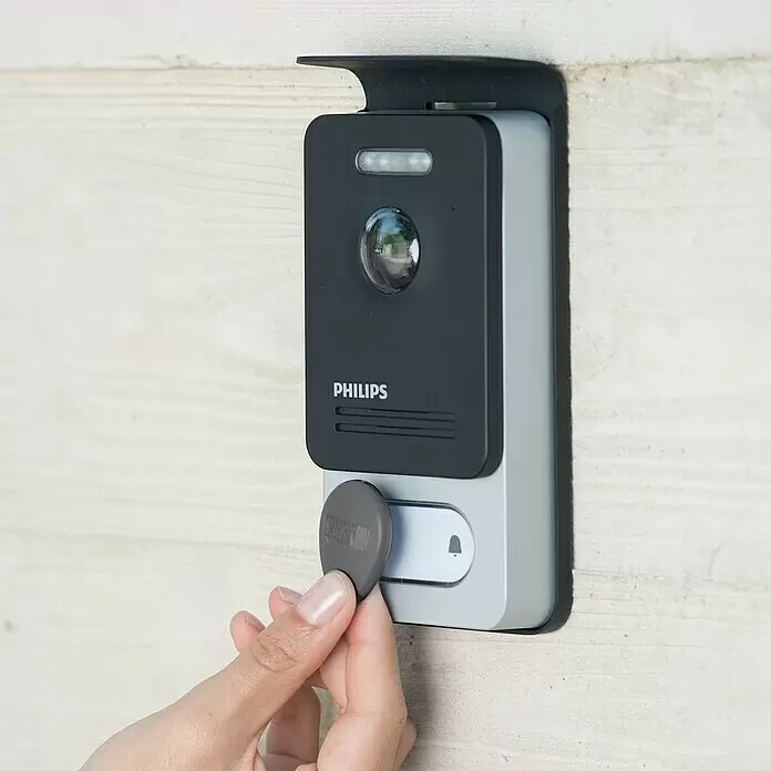 Philips WelcomeEye Videoportero Connect 2 - Plateado - Una mano usa una etiqueta RFID gris en un videoportero Philips negro y plateado montado en una pared de madera clara.