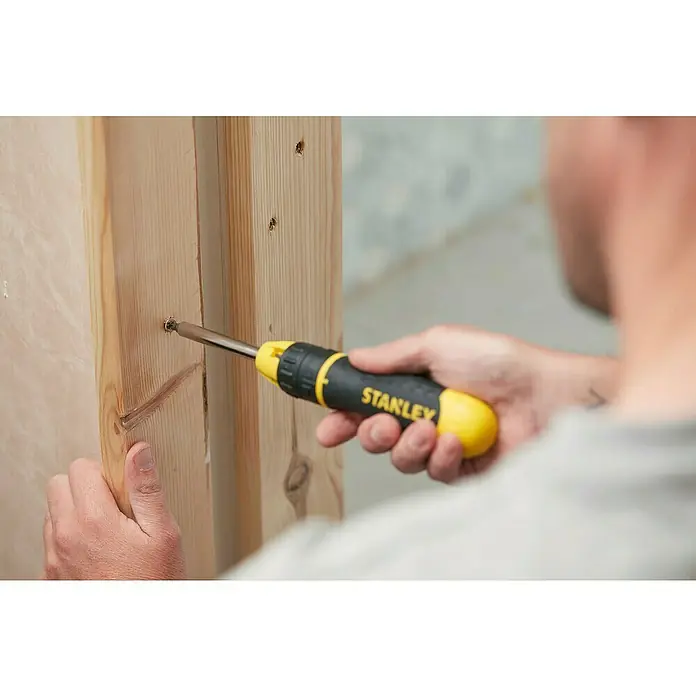 Stanley Destornillador de punta intercambiable 0-68-010 - 10 pzs. - Un artesano atornilla un tornillo en una tabla de madera con un destornillador Stanley amarillo y negro. La herramienta es fácilmente reconocible.