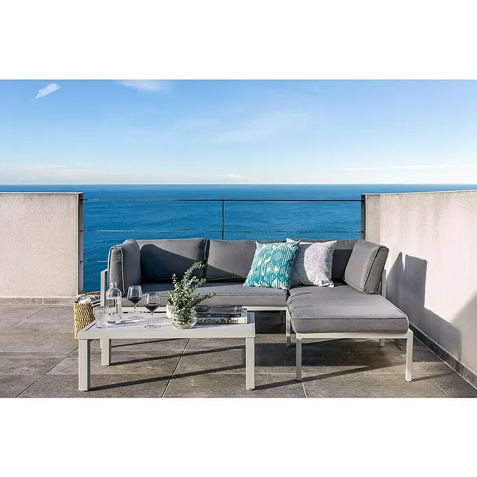 Sunfun Hannah Conjunto de muebles - 3 pzs., Aluminio, Gris claro - Un conjunto de sofá gris con mesa está situado en una terraza con vista al mar. Cojines y complementos decorativos completan la imagen.