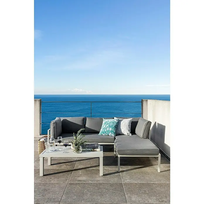 Sunfun Hannah Conjunto de muebles - 3 pzs., Aluminio, Gris claro - Un sofá lounge gris con mesa y cojines en una terraza con vistas al mar. Perfecto para relajarse al aire libre.