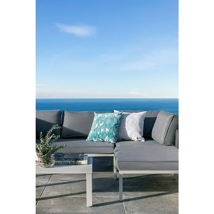 Sunfun Hannah Conjunto de muebles - 3 pzs., Aluminio, Gris claro - Un conjunto de sofá de exterior gris con cojines y una mesa contra un cielo azul. Perfecto para momentos de relajación al aire libre.