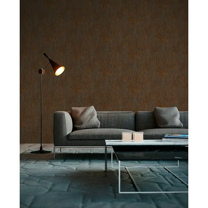 A.S. Création Elements Vliestapete Patina - Braun/Kupfer, Metalloptik, 10,05 x 0,53 m - Wohnzimmer mit einem grauen Sofa, einem Marmortisch und einer Stehlampe, die ein warmes Licht spendet. Die Wand hat eine strukturierte Oberfläche.