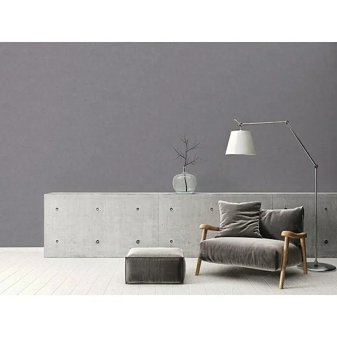 A.S. Création Elements Vliestapete - Dunkelgrau, Uni, 10,05 x 0,53 m - Ein gemütlicher grauer Sessel mit passendem Hocker, ergänzt durch eine elegante Stehlampe und eine moderne Betonkommode.