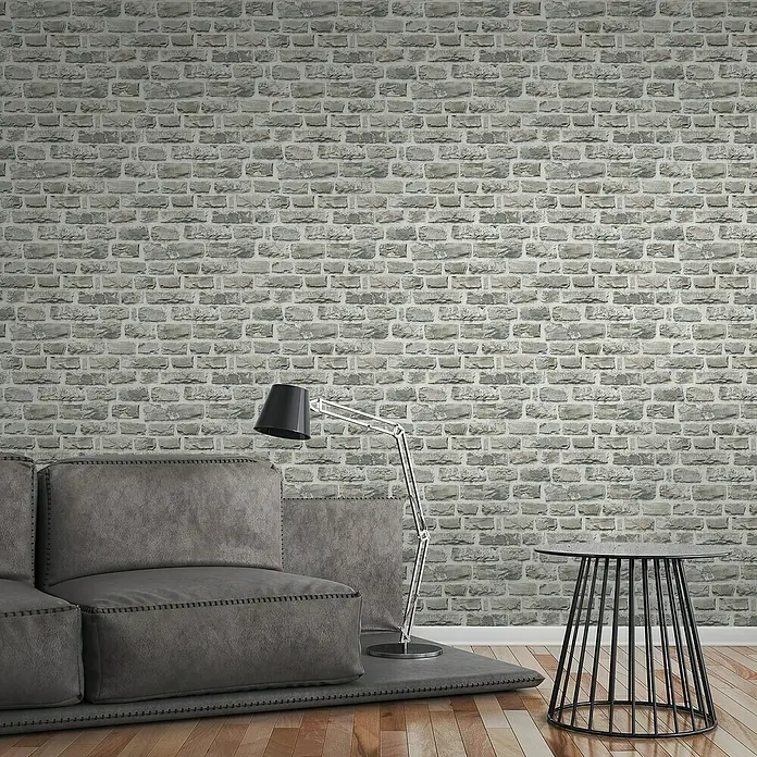 A.S. Création Elements Vliestapete Steine - Creme/Grau, Steinoptik, 10,05 x 0,53 m - Graue Ziegelwand-Tapete als Hintergrund für ein modernes Interieur mit Sofa, Lampe und Beistelltisch.