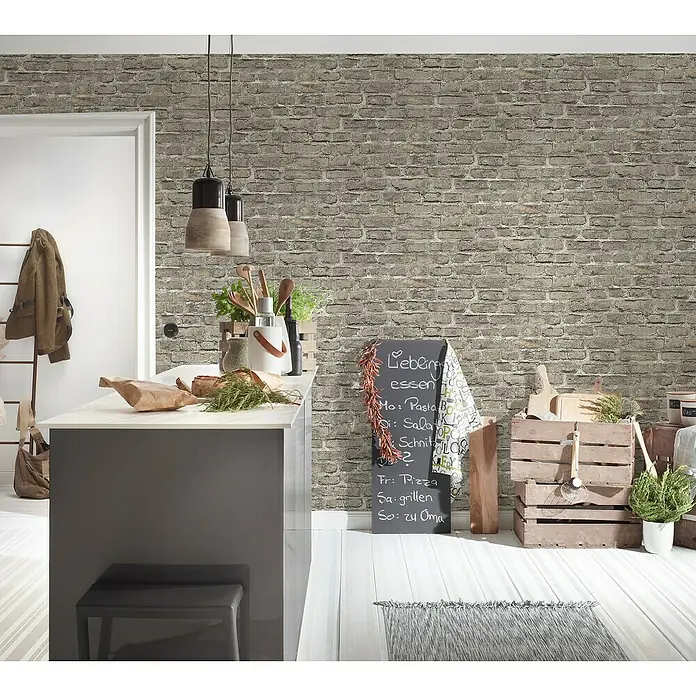 A.S. Création Elements Vliestapete Backsteinmauer - Beige/Grau, Steinoptik, 10,05 x 0,53 m - Ein stilvolles Interieur mit einer Backsteinmauer im Hintergrund, einer modernen Kücheninsel und dekorativen Elementen, die eine gemütliche Atmosphäre schaffen.