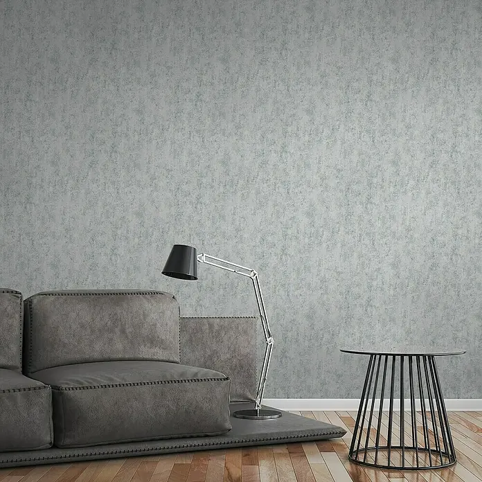 A.S. Création Elements Vliestapete - Hellgrau, Betonoptik, 10,05 x 0,53 m - Einrichtungsszene mit grauer Couch, Stehlampe und Beistelltisch vor einer grauen Wand, die eine moderne Wohnatmosphäre schafft.