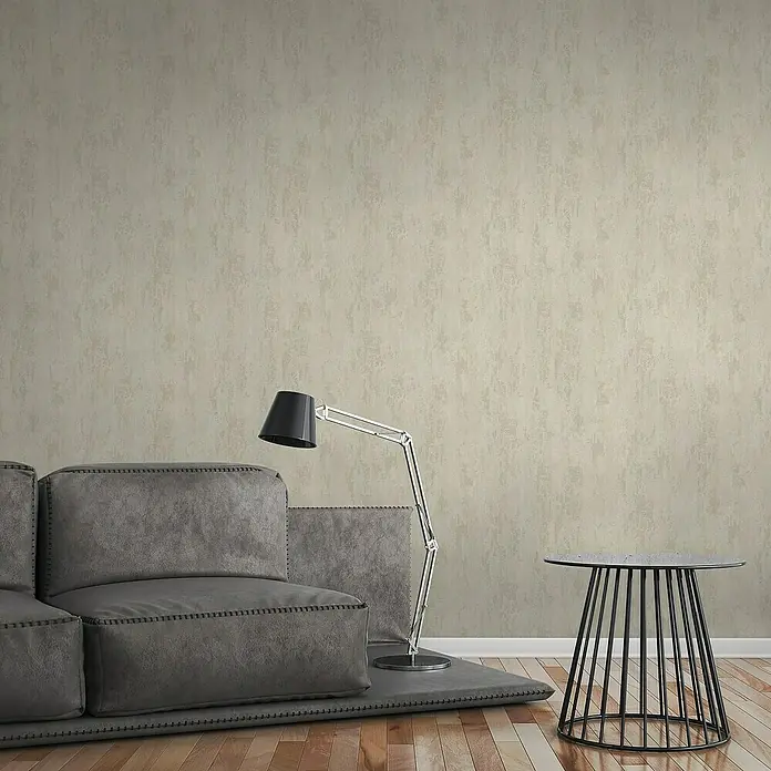 A.S. Création Elements Vliestapete Patina - Beige/Creme, Metalloptik, 10,05 x 0,53 m - Ein graues Ecksofa mit Steppdetails und einer Stehlampe daneben, vor einer strukturierten, hellen Wand. Ein runder Beistelltisch steht daneben.