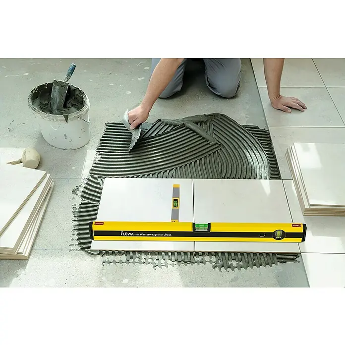 Flüwa Wasserwaage - Länge: 60 cm, Gelb/Schwarz, 16-cm-Lineal - Fliesenleger bei der Arbeit mit einer FLÜWA-Wasserwaage, die auf frisch verlegten Fliesen liegt und die Nivellierung unterstützt. Das Werkzeug gewährleistet Präzision.