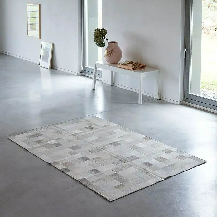 Esbeco Kuhfell Patchwork - Grau, 100 % Echtfell, 180 x 120 cm - Ein modernes Wohnzimmer mit einem Patchwork-Teppich in Grau- und Weißtönen, der auf einem glatten Betonboden liegt.