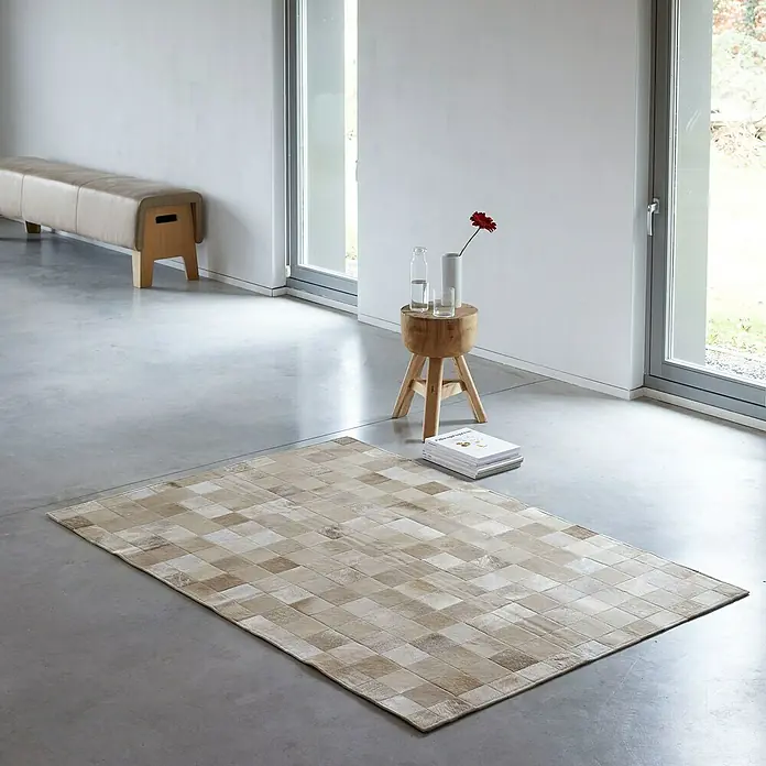 Esbeco Kuhfell Patchwork - Champagner, 100 % Echtfell, 180 x 120 cm - Ein rechteckiger Teppich mit einem geometrischen Muster aus beigen und hellbraunen Quadraten liegt auf einem glatten, grauen Boden.