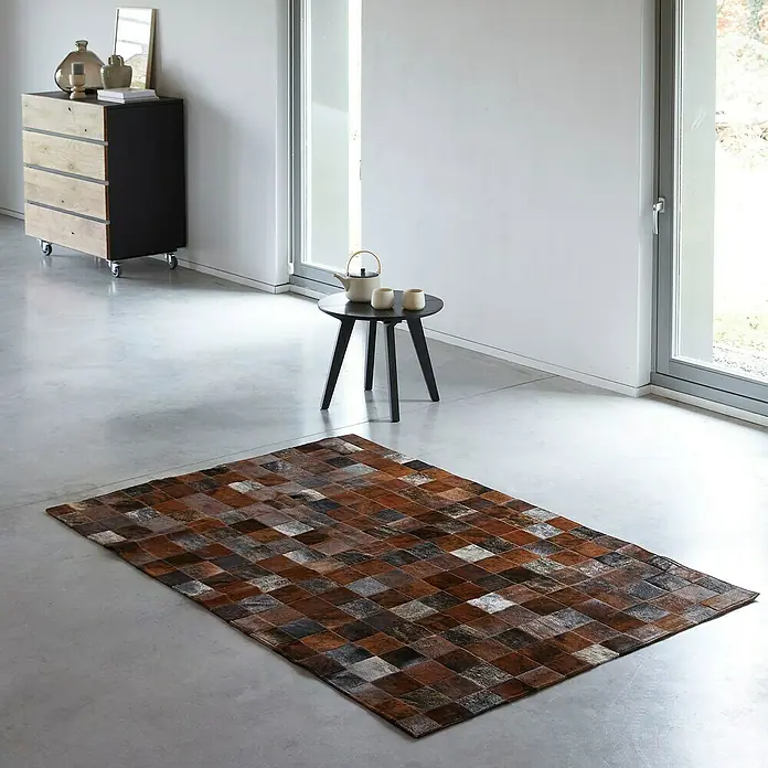 Esbeco Kuhfell Patchwork - Braun, 100 % Echtfell, 180 x 120 cm - Ein Patchwork-Teppich mit braunen und grauen Quadraten liegt auf einem grauen Boden in einem modern eingerichteten Raum.
