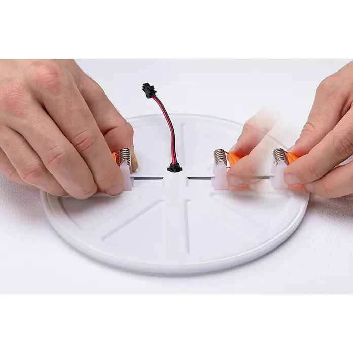 Paulmann Panel LED redondo Veluna - 17 W, Ø x Al: 18,5 x 3,1 cm, Satin, Blanco cálido, No regulable - Primer plano de una lámpara de techo LED que está siendo montada por manos con clips naranjas sobre una superficie blanca.