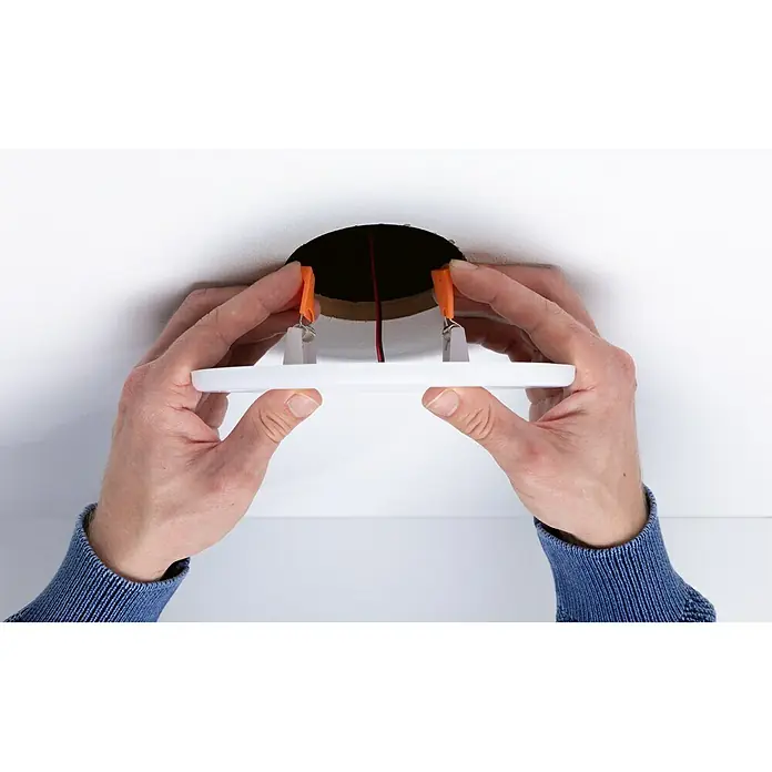 Paulmann Panel LED redondo Veluna - 17 W, Ø x Al: 18,5 x 3,1 cm, Satin, Blanco cálido, No regulable - Una lámpara de techo LED está siendo instalada en una abertura en el techo, sostenida por manos con un suéter azul.