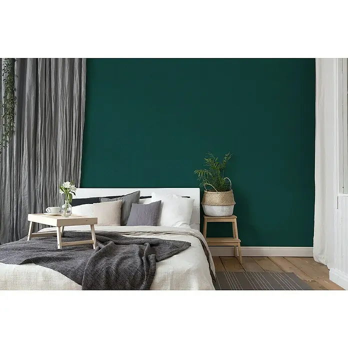 A.S. Création New Elegance Vliestapete - Dunkelgrün, Uni, 10,05 x 0,53 m - Schlafzimmer Interieur mit einem Bett mit Kissen, einer Decke und einem Nachttisch mit Pflanze vor einer smaragdgrünen Wand.