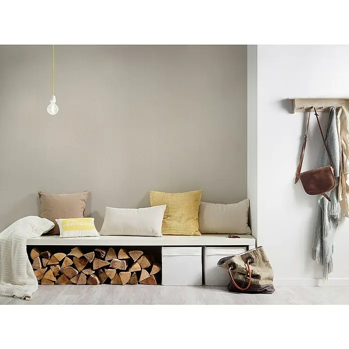 A.S. Création New Elegance Vliestapete - Beige, Uni, 10,05 x 0,53 m - Ein gemütlicher Eingangsbereich mit einer Sitzbank, Kissen und Holzlagerung darunter, ergänzt durch eine Garderobe mit Taschen und Jacken.